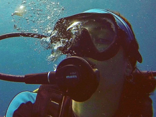 Mitra Nikoo diving underwater with SCUBA diving equipment, wearing dark goggles, a black and blue wet suit, a blue headband, and blowing bubbles through her breathing regulator looking at the camera.