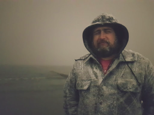 Jameson Clarke standing on an Antarctic beach on a stormy day, ocean waves behind him, wearing a plaid-patterned coat, and grinning at the camera with frost in his beard.