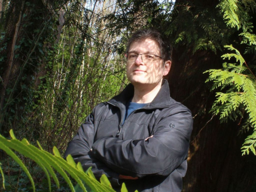 Dr. Julius Csotonyi standing with arms crossed in a forest, partially obscured by a branch of fern leaves, wearing a black windbreaker coat and glasses, and smiling at the camera.