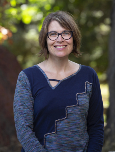Portrait photo of Dr. Amanda Bates standing beneath a tree, wearing dark-rimmed glasses and a long-sleeved shirt with a zigzag pattern across the torso separating two different colours of blue, and smiling at the camera.