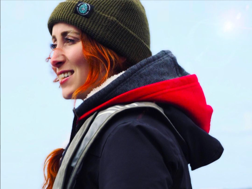 Candid side-view photo of Sydney Dixon bundled in several layers of warm clothing, a green toque, and bright right dyed hair, looking off towards the horizon and smiling.